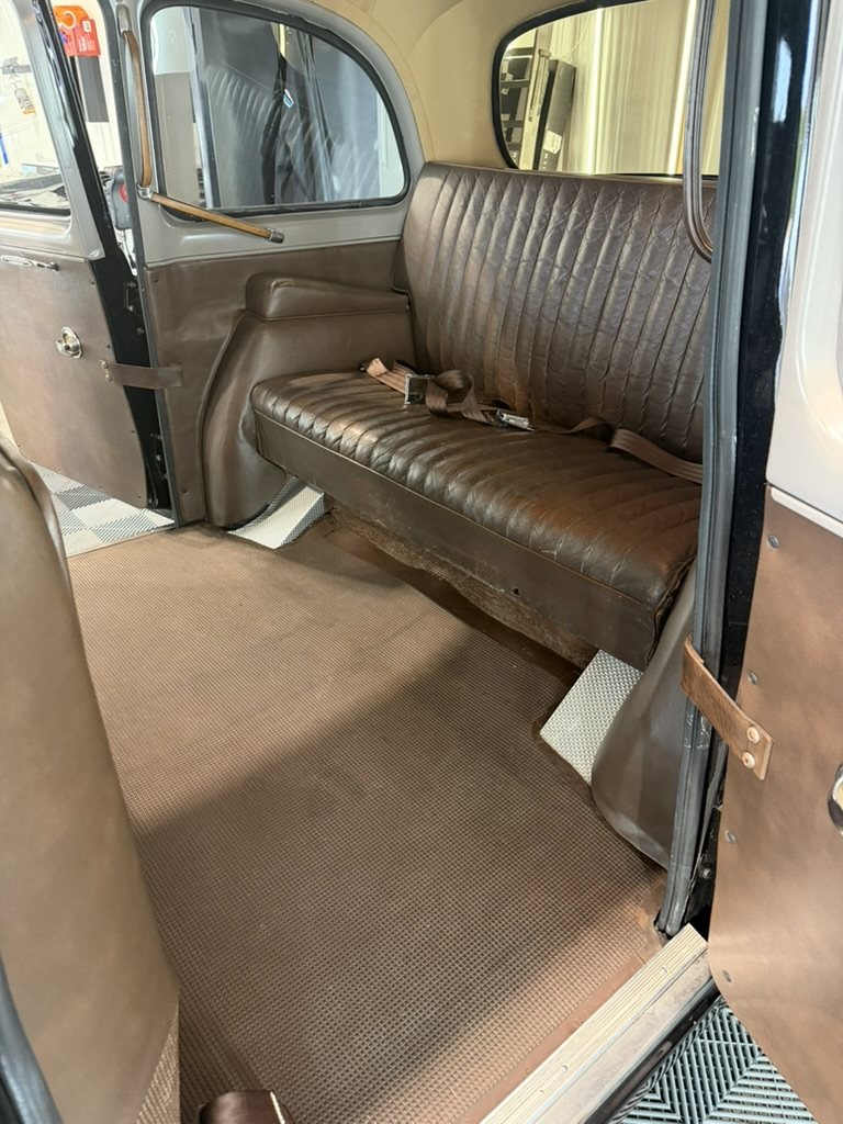 The passenger compartment seen through the open rear door, a pleated brown leather bench seat, brown floor covering and the door's leather pull.