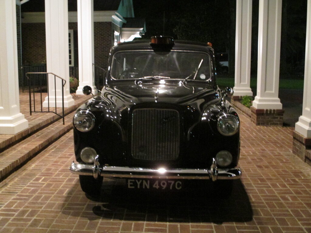The taxi photographed head on at night under a portico, chrome bumper and round headlamps lit, London registration plate EYN 497C.