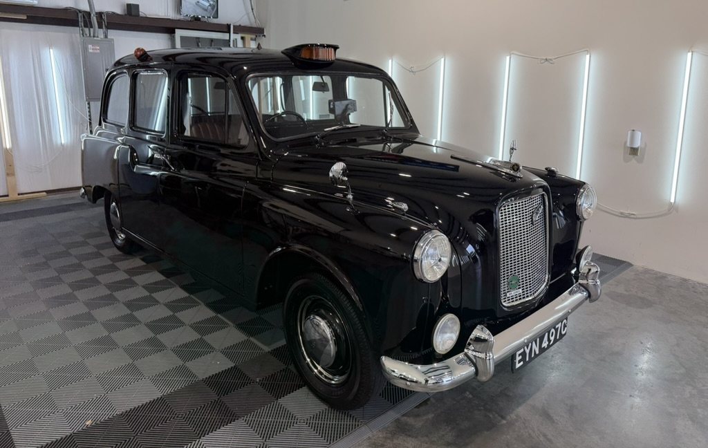 Mr. Jones, a black 1965 Austin FX4 London taxi, freshly detailed and parked on a checkered floor in a detailing shop, paint polished to a mirror, London plate EYN 497C on the chrome front bumper.