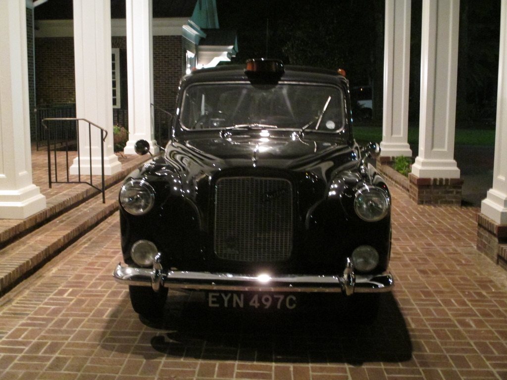 The taxi head on at night beneath a columned porch, amber TAXI roof sign glowing, headlamps and chrome grille catching the light, plate EYN 497C.