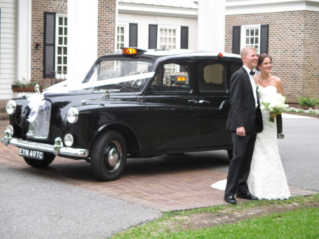 The taxi dressed with white ribbons across the bonnet, parked on a brick drive with a bride and groom standing beside it.